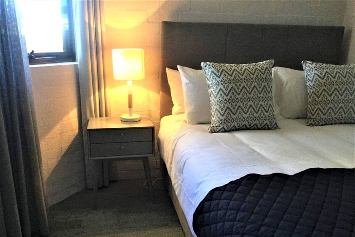 A hotel room with a bed, a bedside table with a lamp, patterned pillows, and a window with curtains.