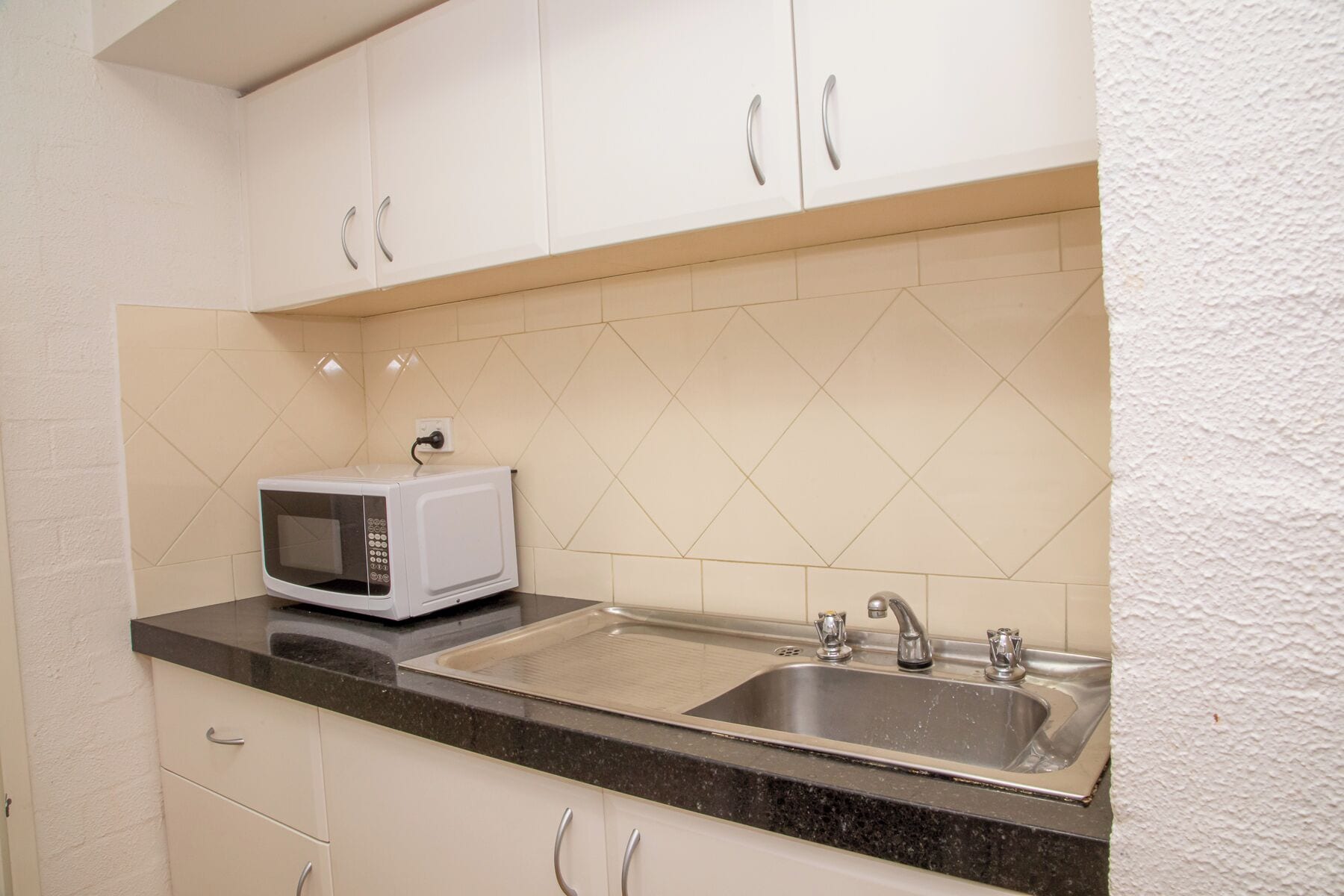 A kitchen with white cabinets, a black countertop, a white microwave, and a stainless steel sink.