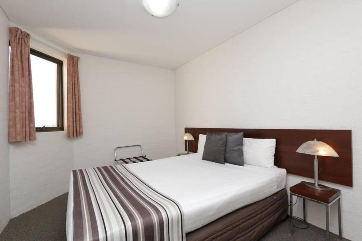 A hotel room with a single bed, a bedside table with a lamp, a window with curtains, and a plain white wall.