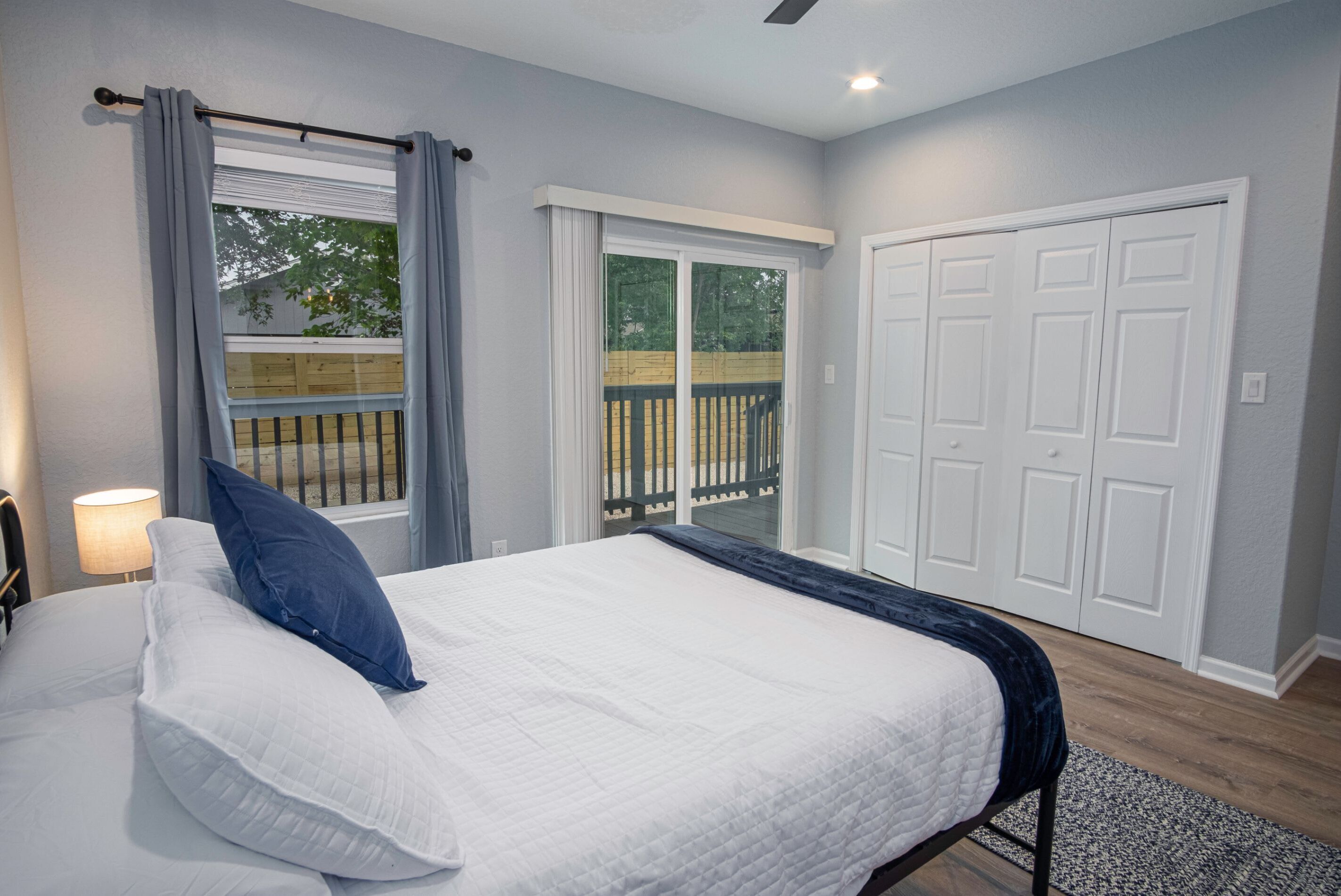 A bedroom with a bed, a nightstand, a window with curtains, a sliding glass door to a balcony, and a white door.