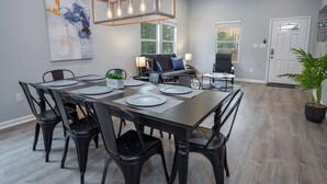 En matplats med ett rektangulärt bord för sex personer, omgivet av svarta stolar. I taket hänger en takfläkt, på väggen sitter en vägglampa och ovanför bordet finns en stor, abstrakt tavla.