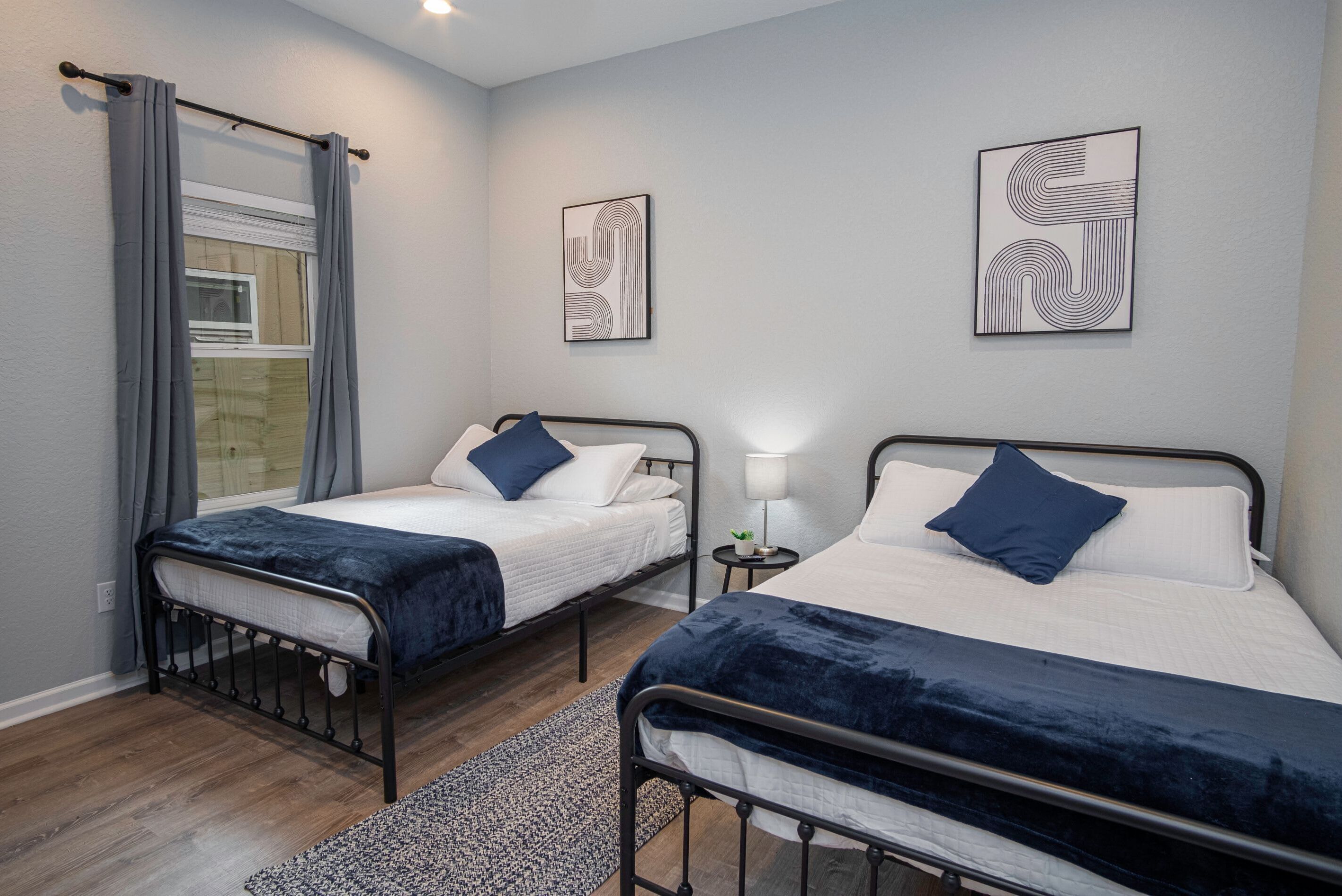 A bedroom with two beds, each with a dark blue headboard and white bedding, a window with grey curtains, and abstract wall art.