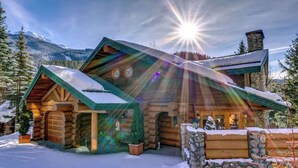 Una cabaña de troncos con chimenea de piedra, techo cubierto de nieve y montañas al fondo.
