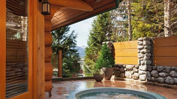 Fachada de madera con un muro de piedra, un jacuzzi y vistas a árboles y montañas.