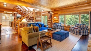 Interior de una cabaña de troncos con escalera, muebles de madera y ventanas amplias.