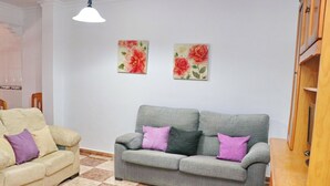 A living room with a grey sofa and a beige sofa, both with purple cushions. There are two floral paintings on the wall and a wooden cabinet to the right. - Cervantes Apartment in Conil (Conil de la Fra)