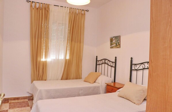 A bedroom with two single beds, a wooden nightstand, a small table, and a window with curtains.