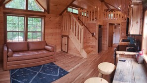 Interior de una cabaña de madera con sofá de cuero, zona de cocina y una escalera.