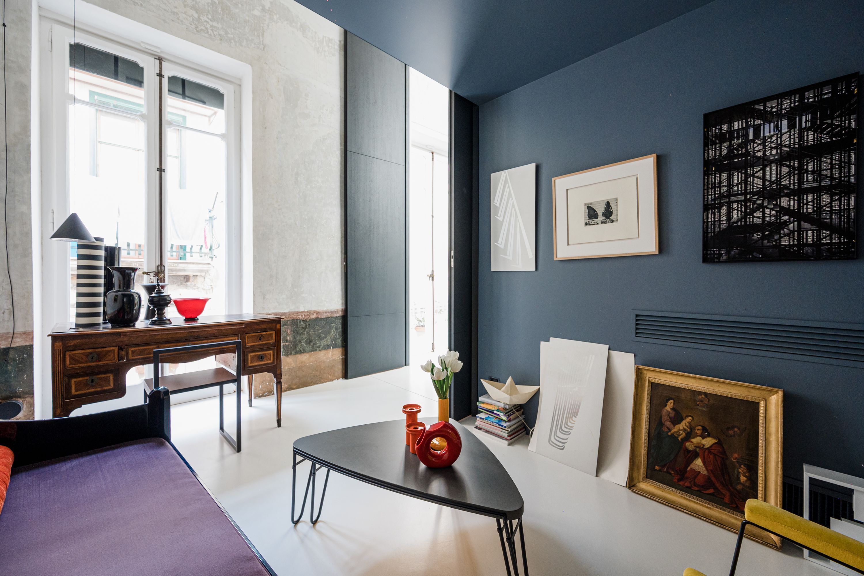 A modern room with a bed, a desk, and a table. There are framed artworks and a vase with flowers on the wall.