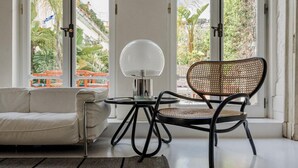 Una sala moderna con un sofá, una silla de mimbre, una mesa redonda y un ventanal con vistas a zonas verdes.