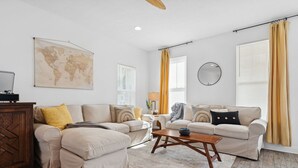 A living room with two beige sofas, a wooden coffee table, a world map on the wall, and a large window with yellow curtains. - Family Getaway•4BR•Games•Fire Pit•Easy Beach Access•Fenced Yard•Pet Friendly  (Panama City Beach)