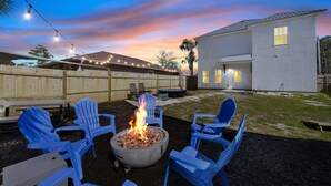An outdoor seating area with blue chairs around a fire pit, a wooden fence, and a house in the background. - Family Getaway•4BR•Games•Fire Pit•Easy Beach Access•Fenced Yard•Pet Friendly  (Panama City Beach)
