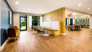 Une pièce spacieuse avec un plancher en bois, une table ronde blanche et plusieurs chaises. Une grande fenêtre offre une vue sur la verdure à l’extérieur.