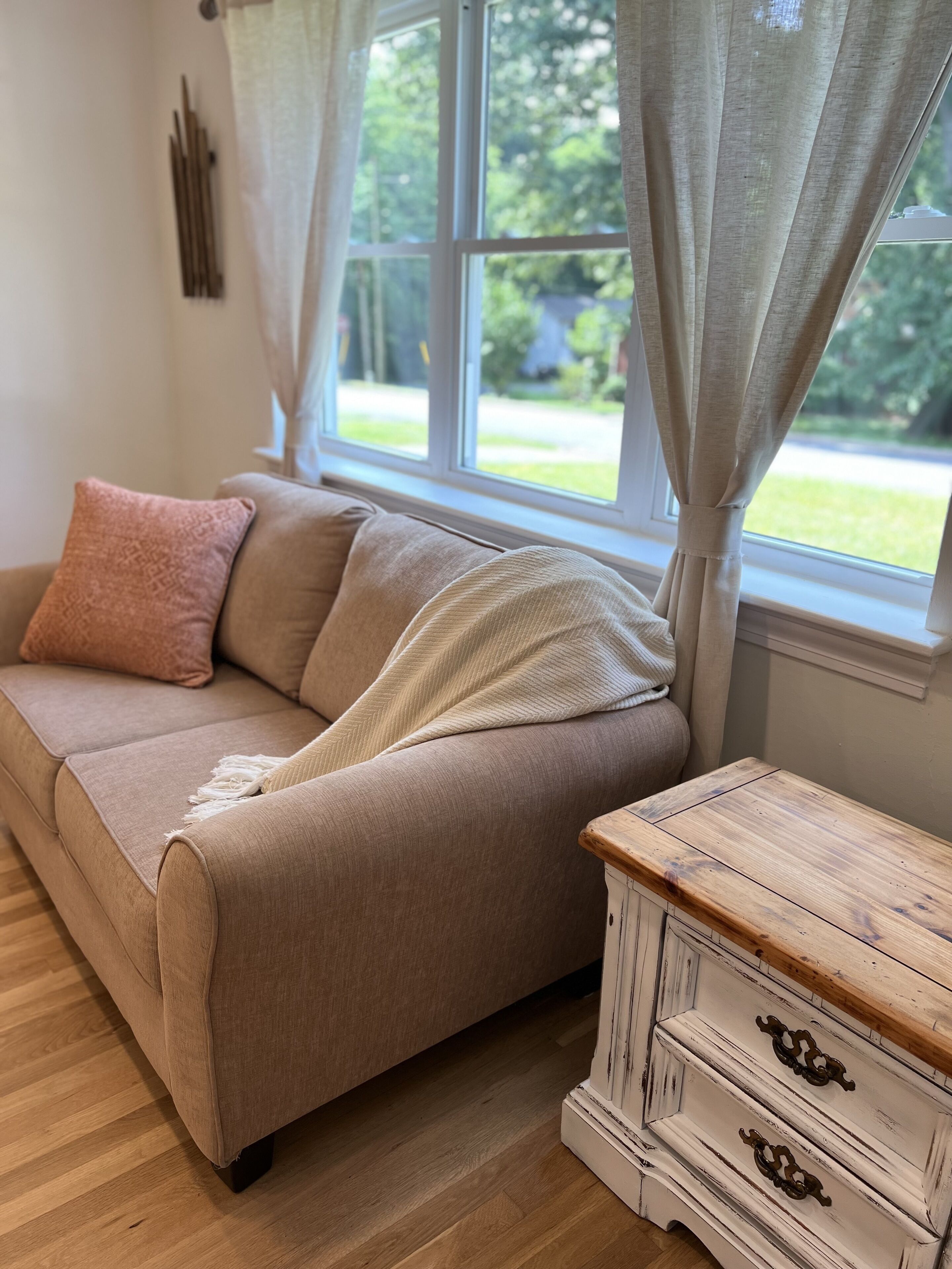 A beige sofa with a throw blanket and a pink pillow, positioned next to a window with curtains. A wooden dresser with drawers is visible to the right.