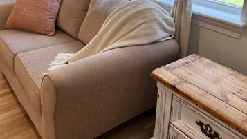 A beige sofa with a throw blanket and a pink pillow, positioned next to a window with curtains. A wooden dresser with drawers is visible to the right.