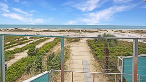 Une vue sur la plage depuis un balcon, avec un ciel dégagé, des sentiers sablonneux et une végétation luxuriante.