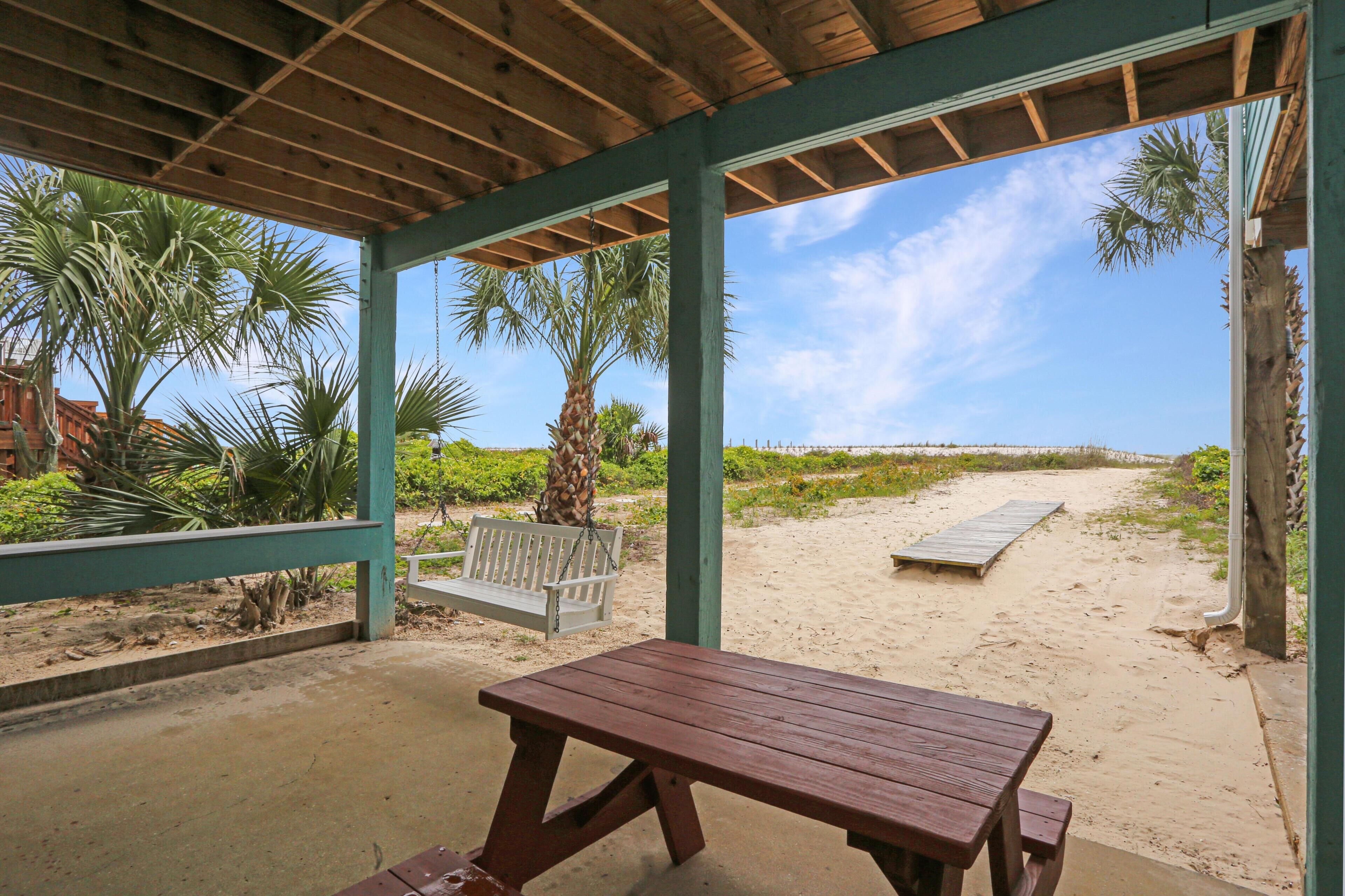 A covered outdoor area with a wooden table and bench, surrounded by palm trees and a sandy path leading to the beach.