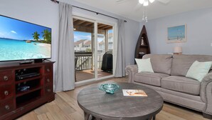 A living room with a TV, a sofa, a coffee table, and a balcony with a view of the beach. - Barrier Dunes 268 - 101 Cape Blessed (Port St. Joe)