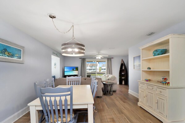 A dining area with a table and chairs, a ceiling fan, a television, and a view of a beach through a window.