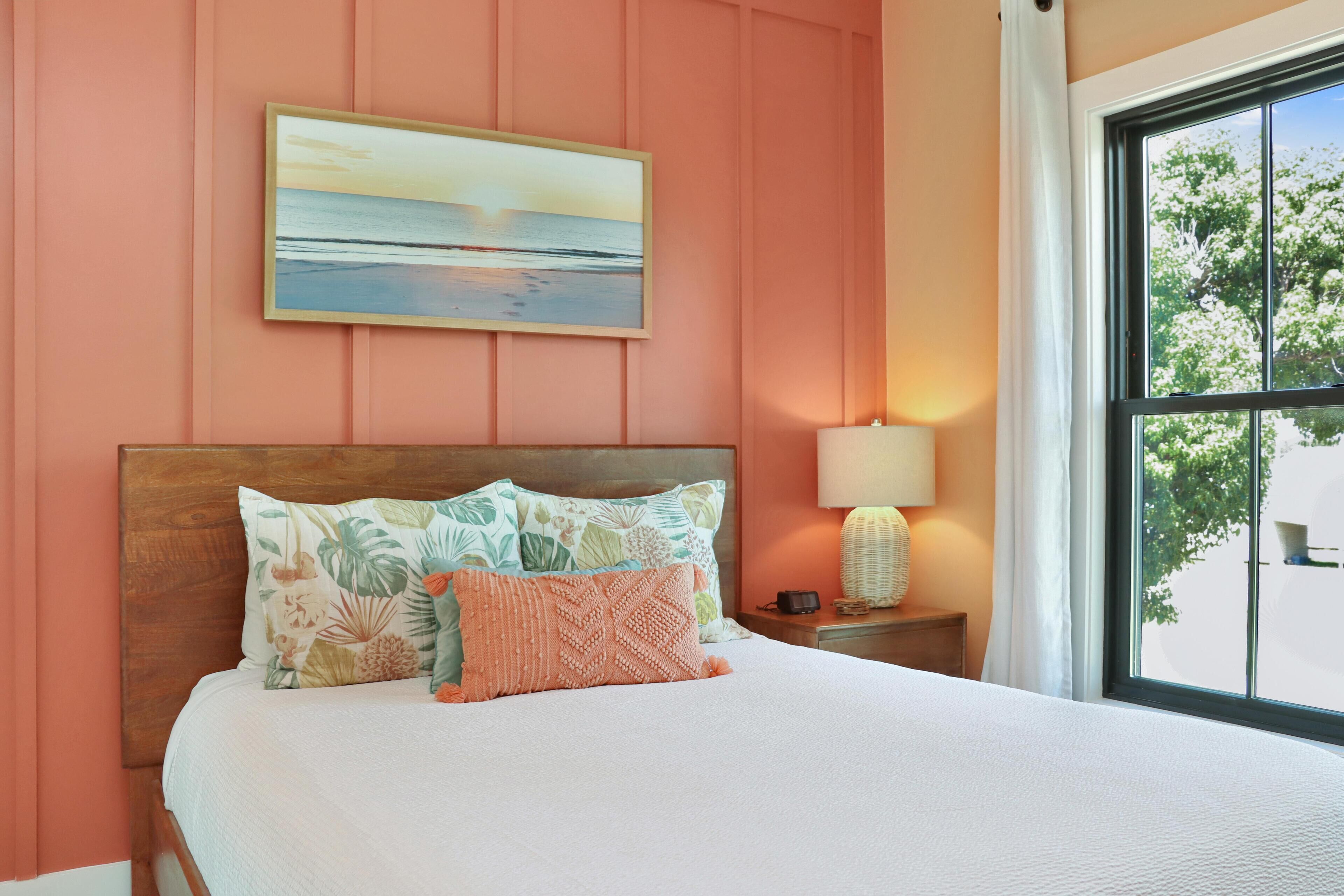 A bedroom with a wooden headboard, a bed with floral and geometric patterned pillows, a nightstand with a lamp, and a painting of a sunset over the ocean.