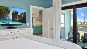 A bedroom with a bed, a TV mounted on the wall, a sliding glass door leading to a balcony, and a view of a kitchen and outdoor area. - Emerald Palms 3 (Port St. Joe)