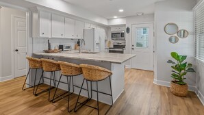 Una cocina moderna con armarios blancos, una isla central con taburetes de bar, electrodomésticos de acero inoxidable y una planta en maceta.