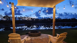 An outdoor patio with two Adirondack chairs, a fire pit, and string lights against a backdrop of a lake and trees. - Paradise Isle (Port St. Joe)