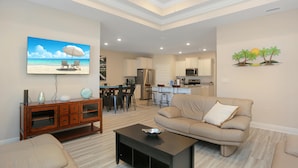 A spacious living room with a beige sofa, a coffee table, and a TV displaying a beach scene. A kitchen is visible in the background with modern appliances. - Paradise Isle (Port St. Joe)