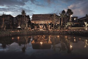 A serene evening scene with a reflective pond, a thatched-roof building, and surrounding vegetation. - Villa Mirah Ubud (Sukawati)