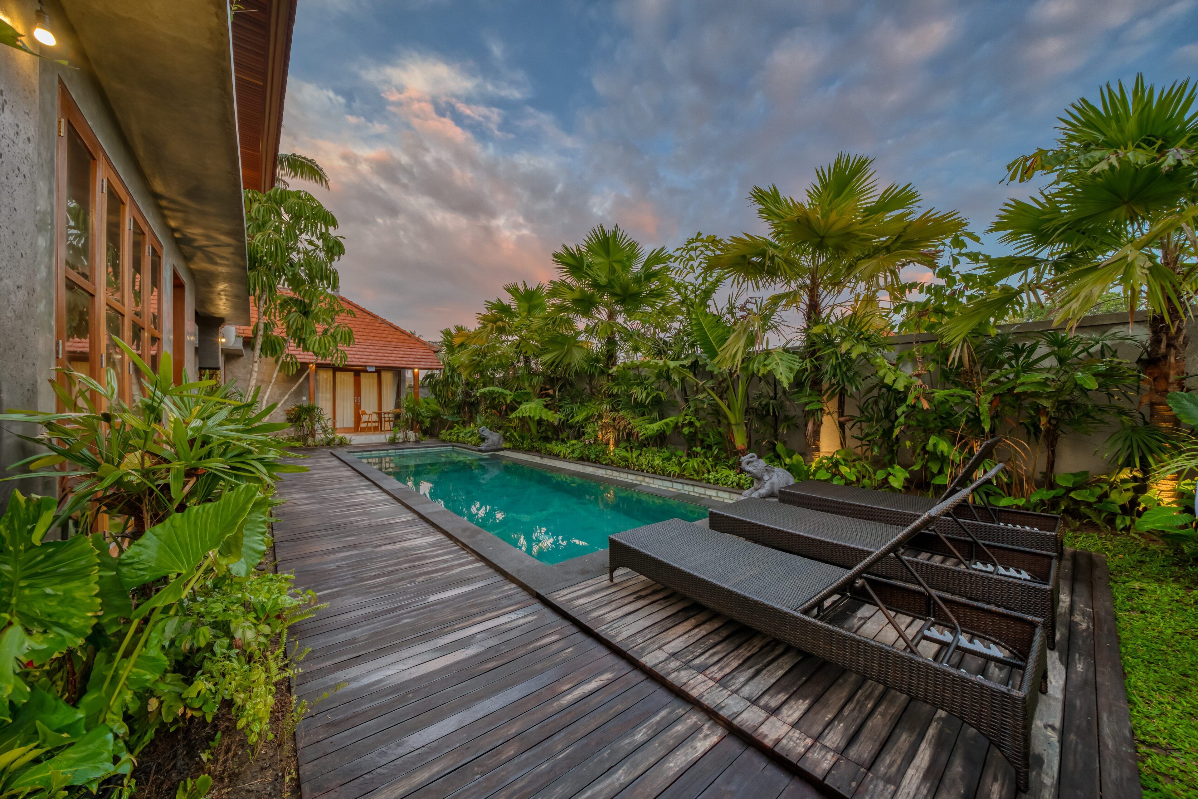 Une terrasse en bois mène à une piscine entourée d’une végétation luxuriante et de plantes tropicales.