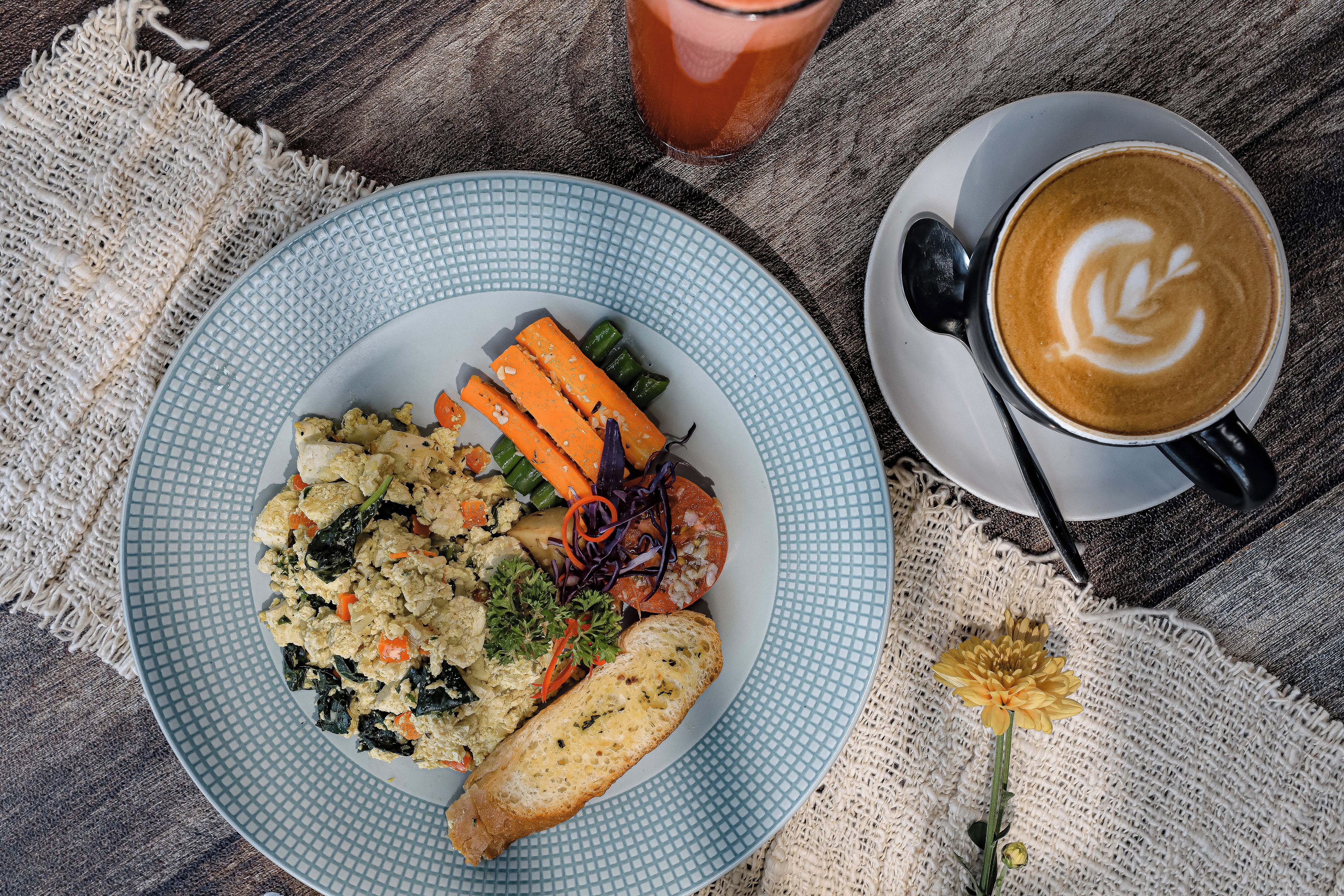Un plato de comida con verduras y una rebanada de pan, una taza de café con arte de latte y un vaso de bebida roja sobre una mesa de madera.