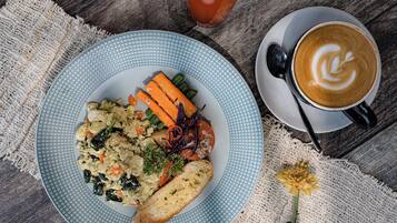 Un plato de comida con verduras y una rebanada de pan, una taza de café con arte de latte y un vaso de bebida roja sobre una mesa de madera.