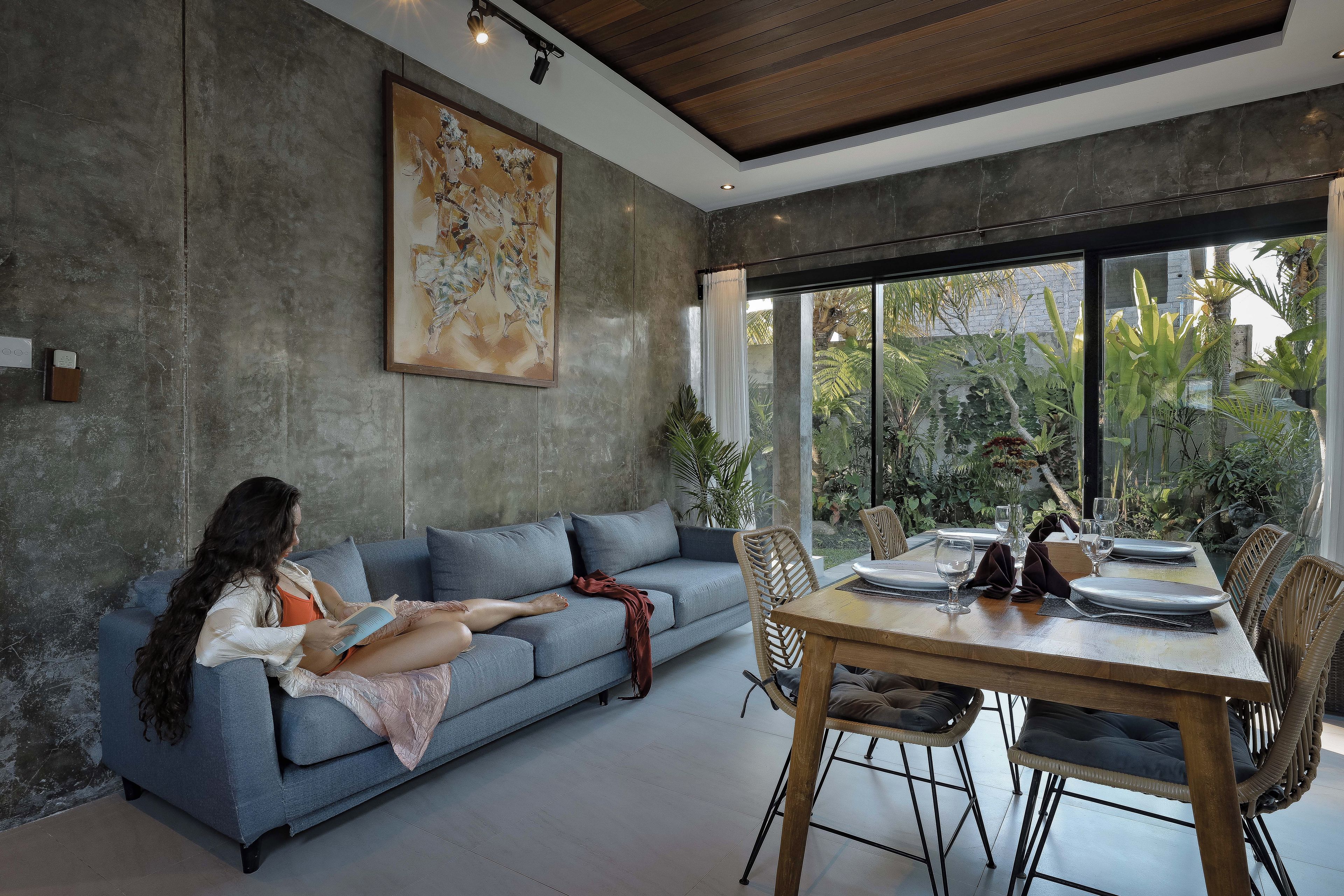 A modern living room with a sofa, a dining table set for two, and a large window with a view of greenery.