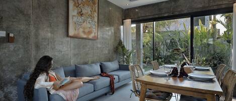 A modern living room with a sofa, a dining table set for two, and a large window with a view of greenery.