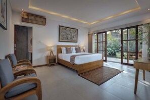 A bedroom with a large bed, a wooden headboard, a nightstand, a bedside table, a chair, and a view of greenery through large windows. - Villa Mirah Ubud (Sukawati)