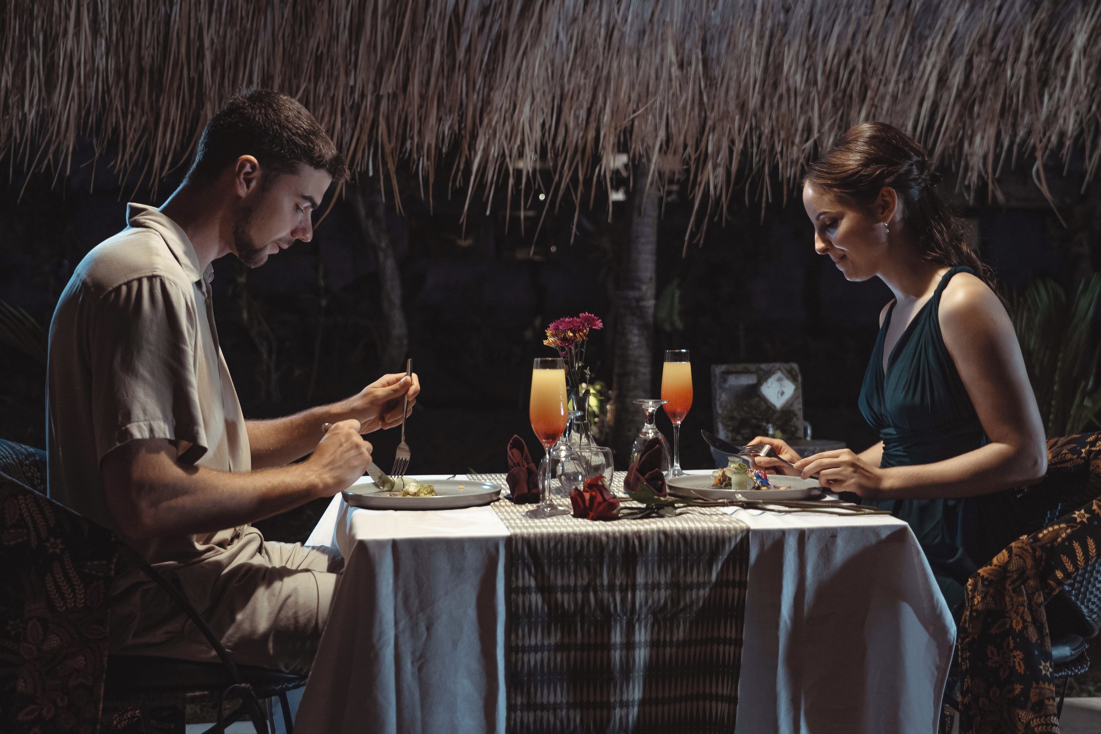 A dining table with a white tablecloth, two people seated, and a thatched roof background.