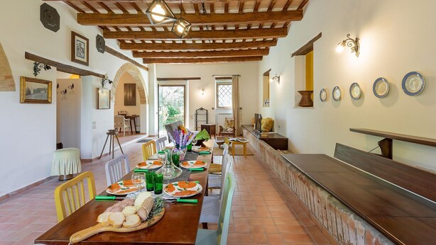Un comedor espacioso con una larga mesa de madera dispuesta para una comida, un banco a lo largo de la pared y un techo rústico con vigas a la vista.