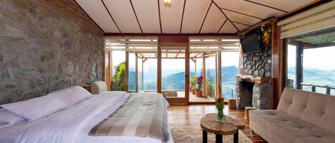 A bedroom with a large bed, a sofa, a stone fireplace, and a view of the mountains.