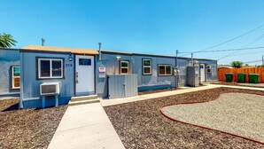 Un bâtiment bleu d’un seul étage, avec des fenêtres numérotées, une caméra de surveillance et une zone délimitée par une clôture, où se trouvent des poubelles vertes.