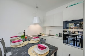 A modern kitchen with white cabinets, a built-in oven, and a dining area with a marble table set for two. - Beachy Vibes- 900m From the Beach (Andora)