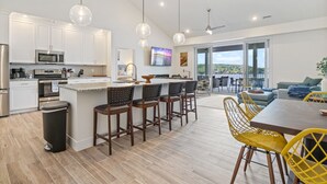 A modern kitchen with white cabinets, a central island, and bar stools. There is a dining area with a table and yellow chairs.