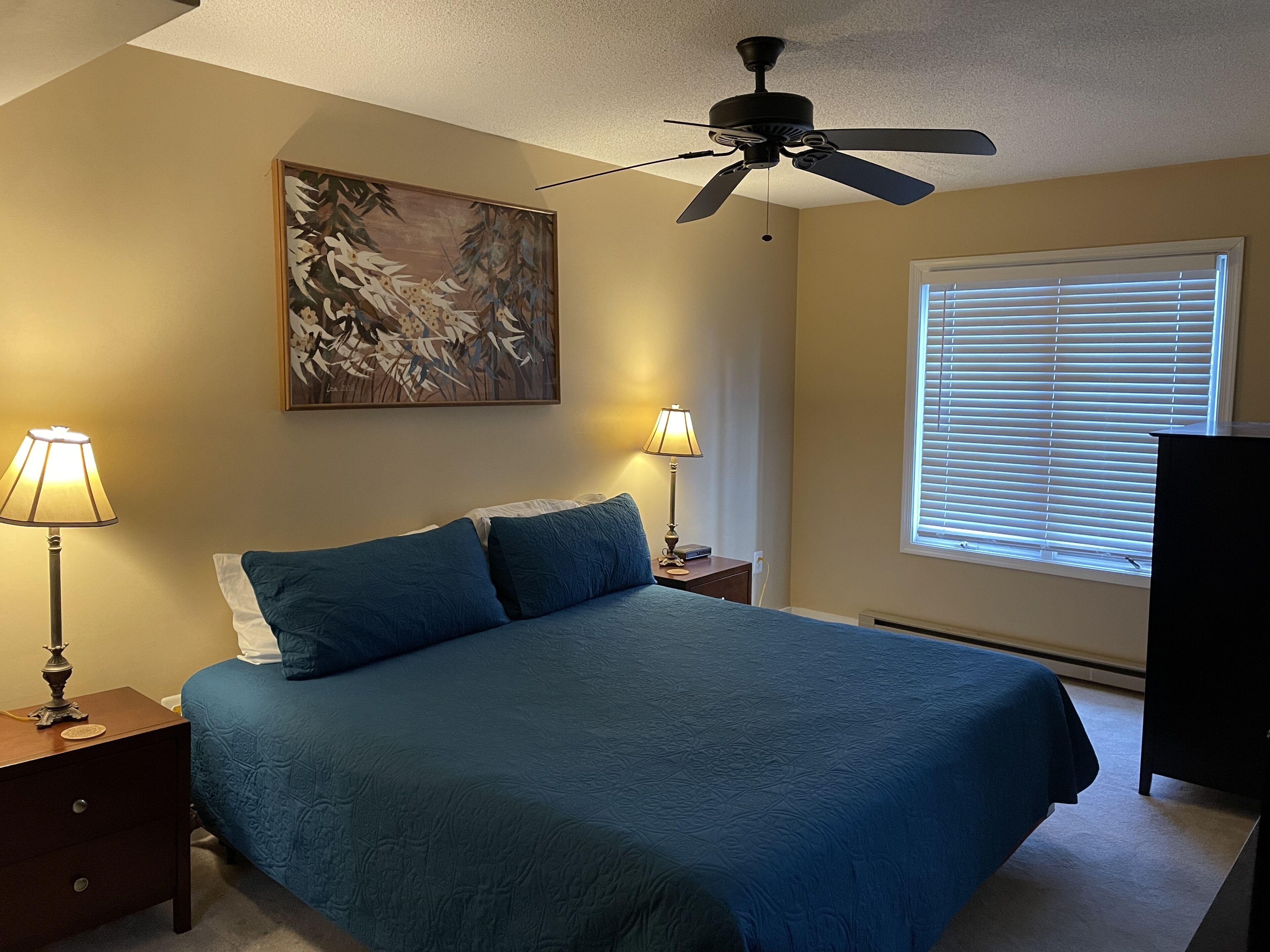 A bedroom with a bed, two bedside lamps, a ceiling fan, a window with blinds, and a painting on the wall.