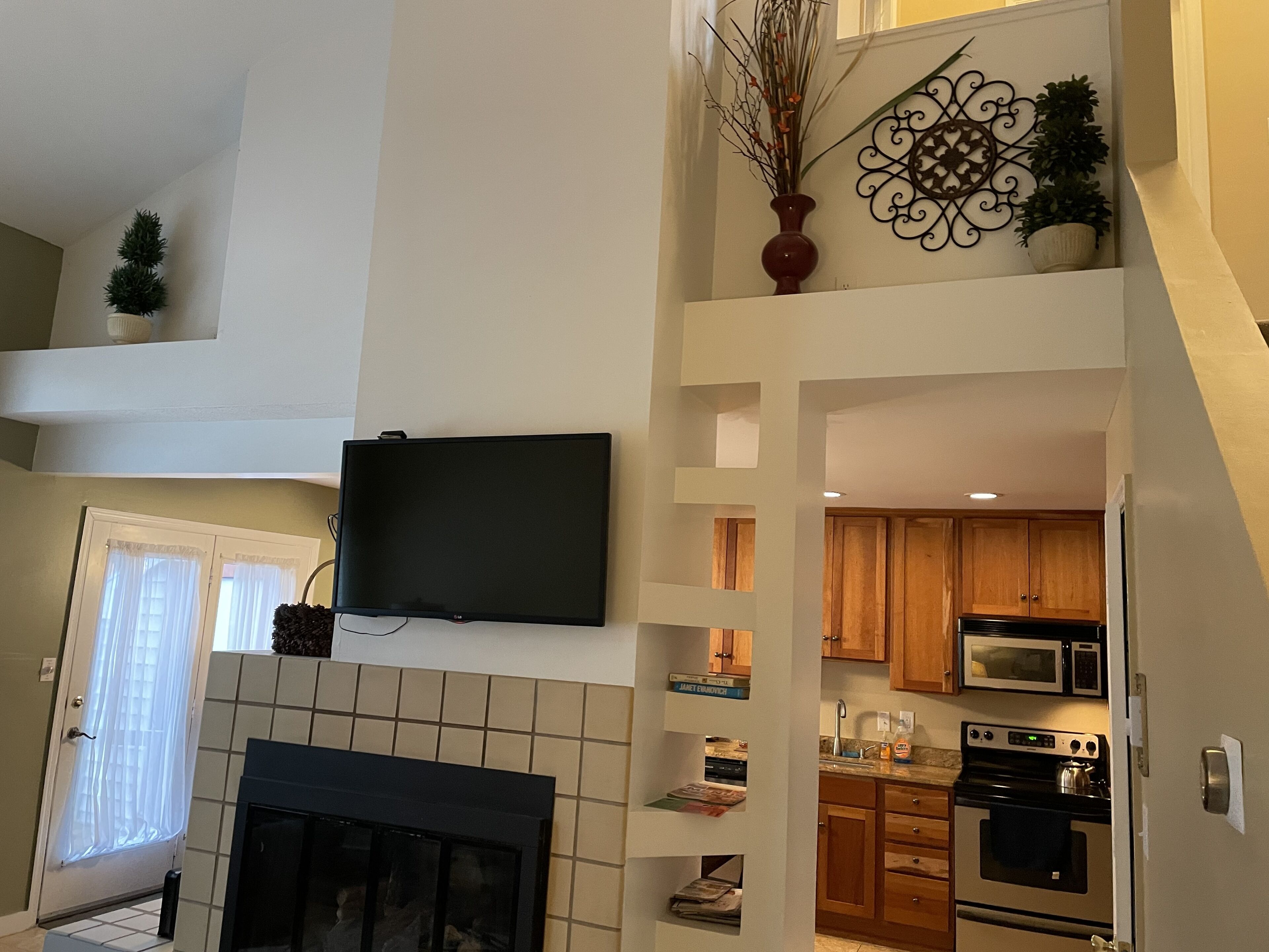 A modern kitchen with a flat-screen TV mounted above the fireplace, wooden cabinets, and a microwave.