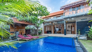 A swimming pool with lounge chairs, a covered patio, and a two-story building with a tiled roof. - Villa Padma Nusa Dua (Nusa Dua)