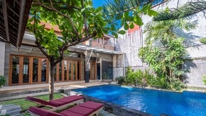 A swimming pool with lounge chairs, a tree, and a building with large windows and a balcony. - Villa Padma Nusa Dua (Nusa Dua)