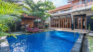 A swimming pool with lounge chairs, a covered patio, and a two-story building with large windows and a balcony. - Villa Padma Nusa Dua (Nusa Dua)