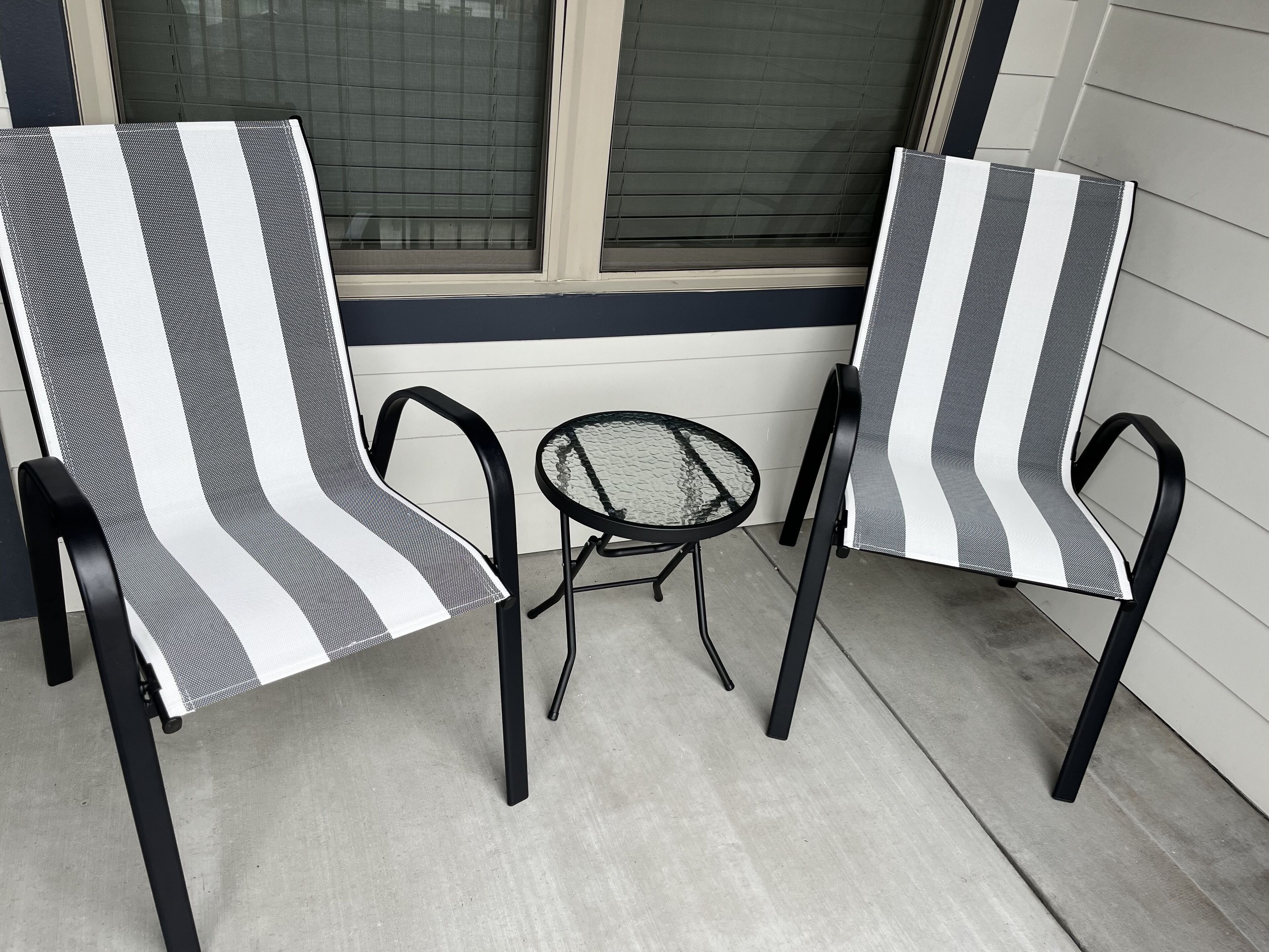 Deux chaises dotées d’un revêtement en tissu à rayures et d’une structure en métal noir, placées sur une terrasse en béton, à côté d’une fenêtre munie de stores.