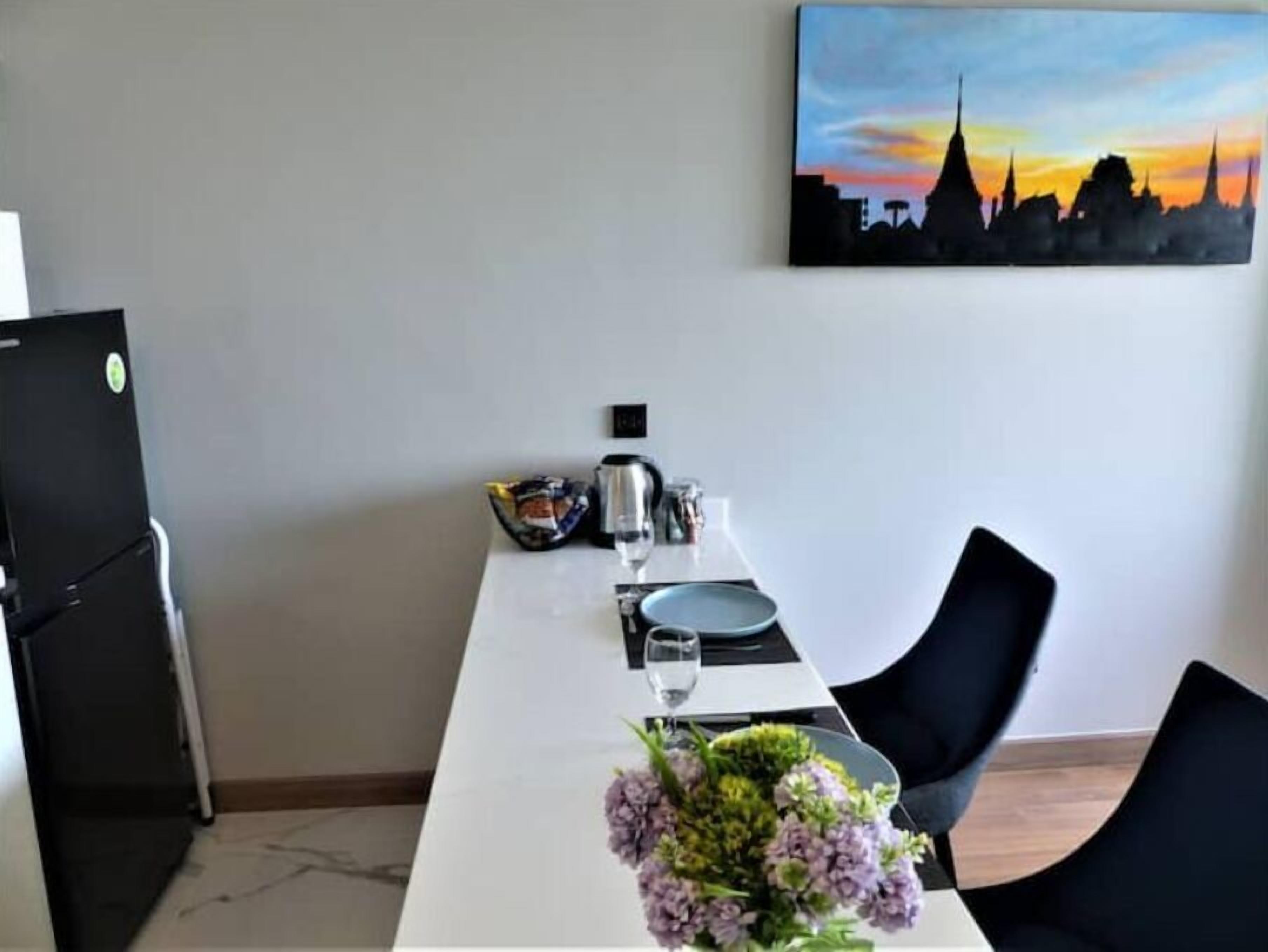A dining area with a white table set for two, a vase of flowers, a painting of a city skyline, and a black refrigerator.