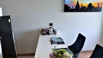 A dining area with a white table set for two, a vase of flowers, a painting of a city skyline, and a black refrigerator.