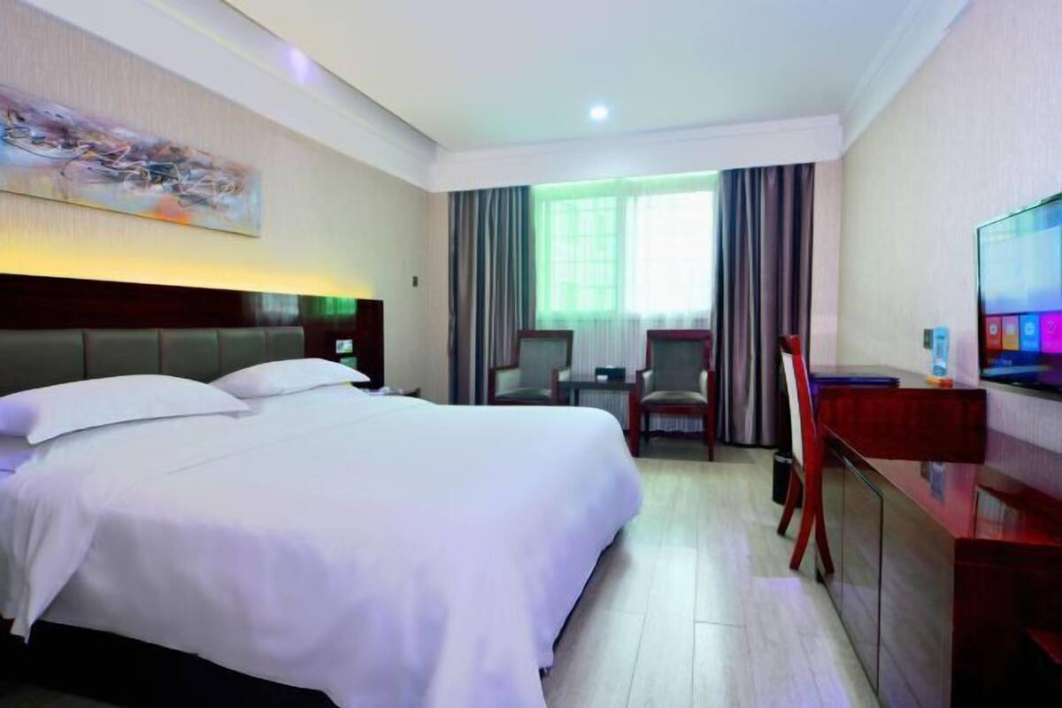 Une chambre d’hôtel avec un grand lit, un bureau, une chaise, une télévision et une fenêtre avec des rideaux.